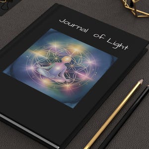 May include: Black journal with the text "Journal of Light" and an illustration of a person meditating within a geometric design. The cover art features a colorful, ethereal design. Gold and black pencils and gold binder clips are also visible.