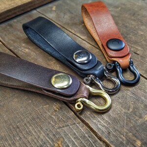 May include: Three leather keychains with different colored straps and metal hooks. The keychain on the left has a brown strap and a gold hook. The keychain in the middle has a black strap and a silver hook. The keychain on the right has a brown strap and a black hook.