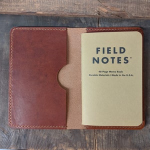 May include: A brown leather notebook cover with a tan interior and a Field Notes memo book. The memo book is tan with black text that reads "FIELD NOTES" and "48-Page Memo Book Durable Materials / Made in the U.S.A."