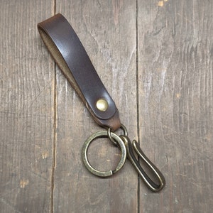 May include: A brown leather key chain with a brass key ring and a brass hook. The key chain is on a wooden surface.