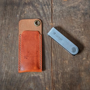 May include: An open, orange leather case with a brass grommet and a silver Ledger device. The case is stitched with light-colored thread. The Ledger device is silver with the word "Ledger" printed on it.