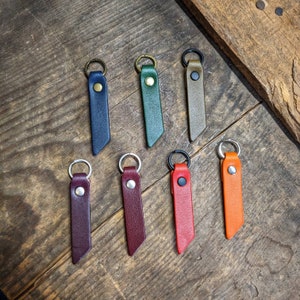 May include: A collection of leather keychains in various colors, including navy blue, green, and orange. Each keychain features a metal ring and a snap closure. The keychains are arranged on a rustic wooden surface.