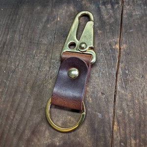 May include: A brown leather key chain with a gold metal hook and a gold metal ring. The key chain is on a wooden surface.