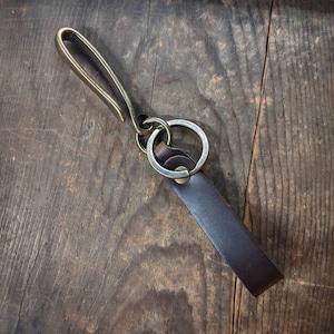 May include: A leather key fob with a brass hook and ring. The dark brown leather strap is attached to a brass ring, which is connected to a brass hook. The key fob is shown on a rustic wooden surface.