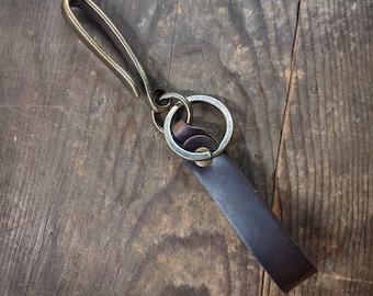 Op de afbeelding: Een leren sleutelhanger met een messing haak en ring. De donkerbruine leren band is bevestigd aan een messing ring, die is verbonden met een messing haak. De sleutelhanger wordt getoond op een rustiek houten oppervlak.