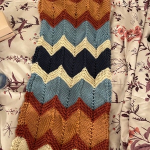 K&ouml;nnte beinhalten: Eine gestrickte Decke mit Chevron-Muster in den Farben Creme, Blau, Braun und Rost.
