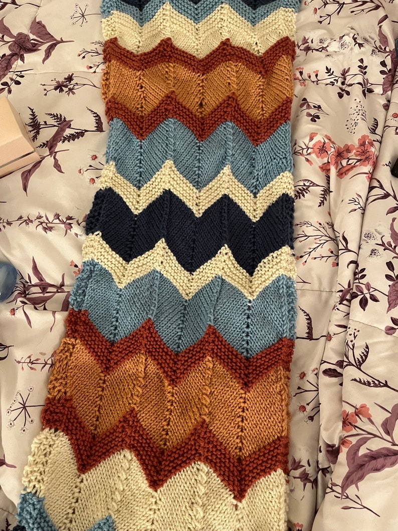 K&ouml;nnte beinhalten: Eine gestrickte Decke mit Chevron-Muster in den Farben Creme, Blau, Braun und Rost.
