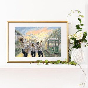 May include: A framed print depicts a jazz band playing in front of a green streetcar. The musicians wear white shirts, black pants, and hats. The background features a colorful sunset and buildings, with a vase of white flowers and greenery in the foreground.