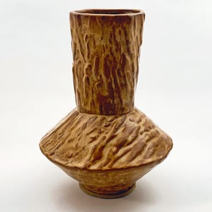 Könnte beinhalten: Eine Keramikvase mit brauner Glasur und einer strukturierten Oberfläche, die an Baumrinde erinnert. Die Vase hat einen breiten, ausgestellten Boden und einen schmalen Hals.