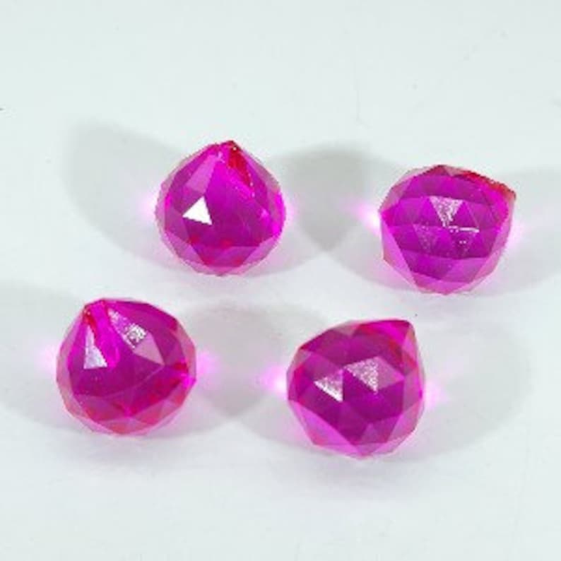 May include: Four faceted, magenta-colored crystal spheres. Each sphere has a complex, geometric design with multiple flat surfaces that reflect light. The crystals are arranged on a white surface, with subtle shadows adding depth.