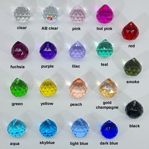 May include: A collection of faceted crystal pendants in various colors, including clear, pink, red, purple, green, yellow, and blue. Each pendant has a small hole for hanging and is labeled with its color name.