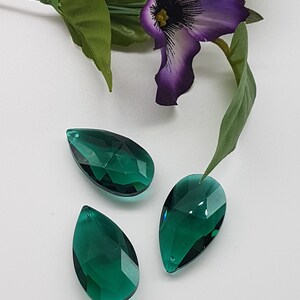 May include: Three teardrop-shaped crystal prisms in a deep emerald green color. The prisms are faceted and reflect light.