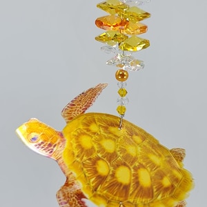 May include: A yellow and orange crystal suncatcher with a turtle design. The suncatcher has a crystal teardrop hanging from the bottom.