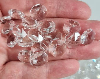 May include: A close-up of a hand holding a pile of clear crystal beads. The beads are faceted and have a hole in the center. They are perfect for making jewelry or adding sparkle to crafts.