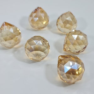 May include: Six faceted, amber-colored crystal pendants and spheres are arranged on a white surface. The crystals have a geometric design, reflecting light. The shapes include teardrop and spherical forms, creating a decorative display.