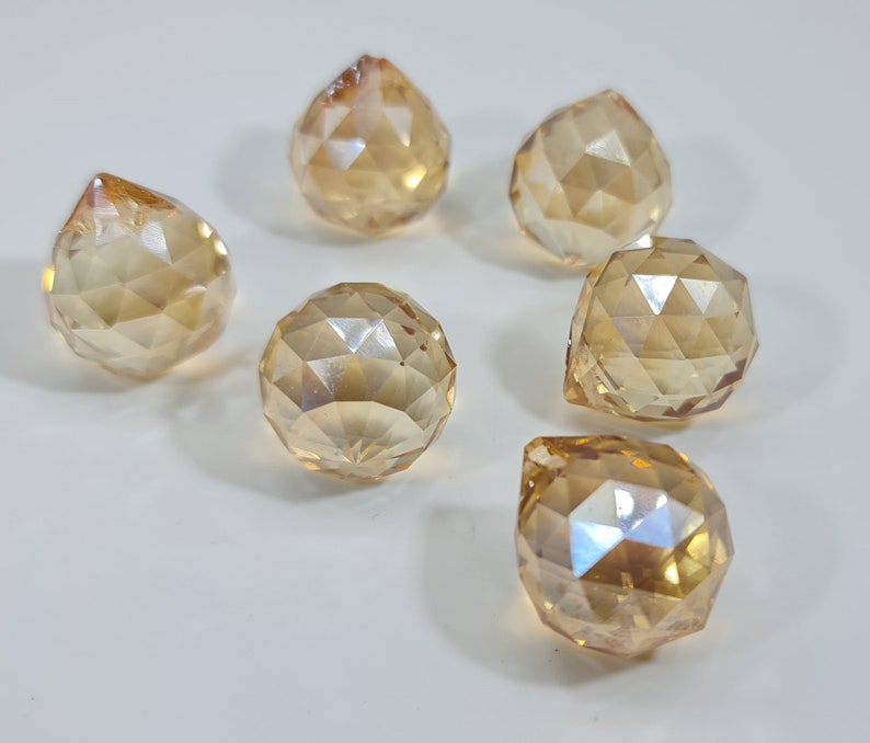 May include: Six faceted, amber-colored crystal pendants and spheres are arranged on a white surface. The crystals have a geometric design, reflecting light. The shapes include teardrop and spherical forms, creating a decorative display.