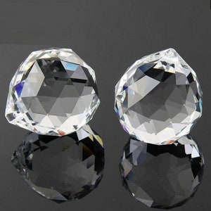 May include: Two clear crystal ball ornaments with faceted surfaces. The ornaments are round and have a slight teardrop shape. They are reflecting light and creating rainbows.