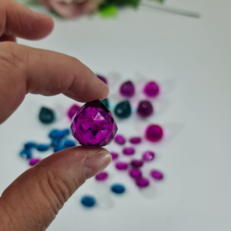 May include: A faceted, teardrop-shaped purple crystal bead held between fingers, with a collection of various small, colorful, translucent crystal beads in shades of purple, blue, and teal scattered in the background.