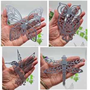 Peut inclure: Quatre ornements en métal argenté découpés : un papillon, un hibou, un colibri et une libellule. Chaque ornement présente des détails découpés complexes. Le papillon a des motifs d'ailes détaillés, tandis que le hibou a un corps détaillé. Le colibri et la libellule ont des ailes détaillées.