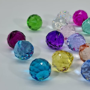 May include: A collection of faceted crystal balls in various colors, including green, purple, blue, and yellow. The spheres are arranged on a white surface, reflecting light and creating a sparkling effect. These decorative orbs could be used for crafts or home decor.