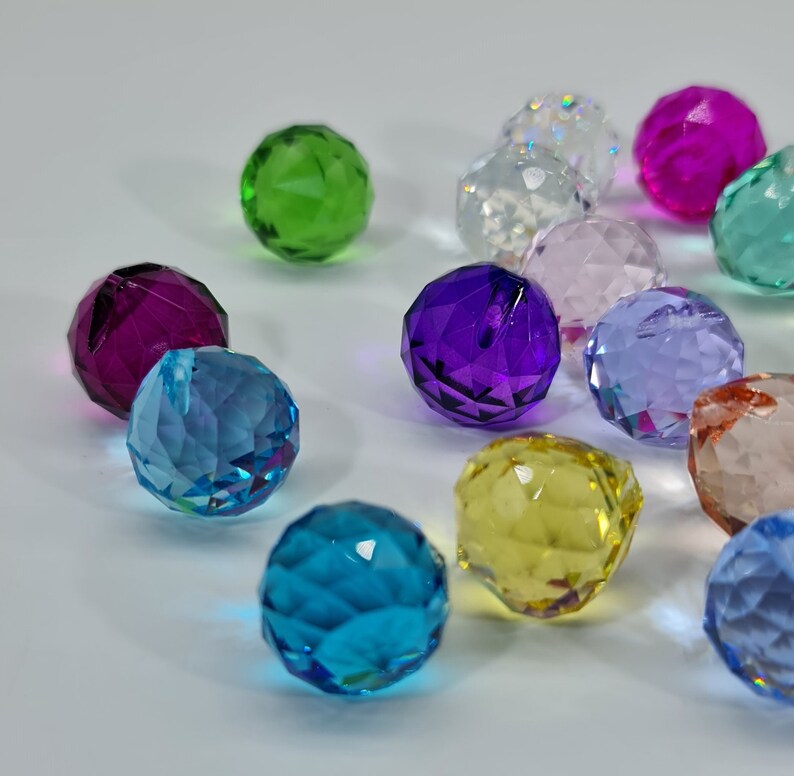 May include: A collection of faceted crystal balls in various colors, including green, purple, blue, and yellow. The spheres are arranged on a white surface, reflecting light and creating a sparkling effect. These decorative orbs could be used for crafts or home decor.