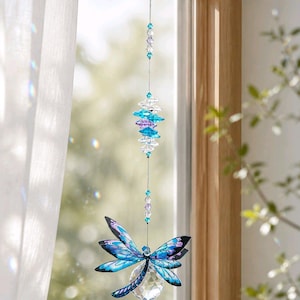 Puede incluir: Un atrapasoles colgante con un adorno de libélula con alas estampadas en azul y morado. La libélula está suspendida por un hilo fino adornado con cuentas de cristal facetado transparentes, azul claro y moradas y una gran esfera de cristal.