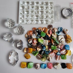 May include: Assortment of colourful iridescent shell beads and clear glass beads in various shapes and sizes. The beads are arranged on a white surface. There is also a clear plastic container with small clear glass beads.