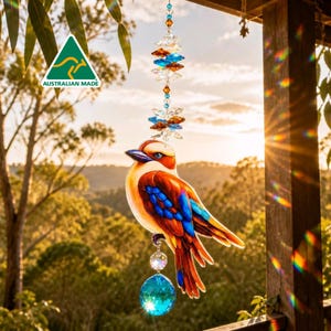 Puede incluir: Un atrapasoles colgante con una ilustración colorida de un kookaburra con plumas marrones, naranjas y azules. El ave está suspendida de una cadena vertical de cristales transparentes, azules y ámbar, con una gran esfera de cristal azul facetado.