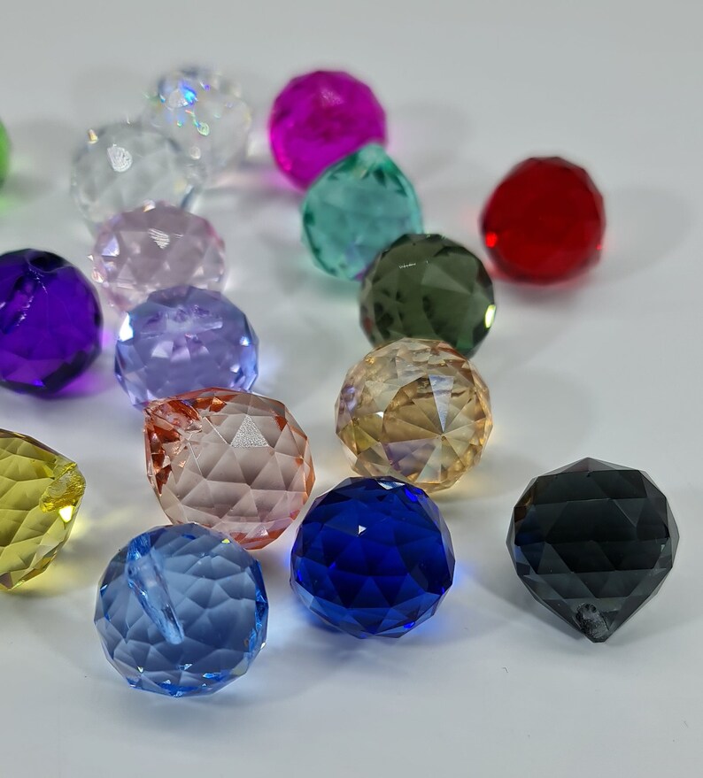 May include: A collection of multi-colored, faceted crystal beads in various hues including red, blue, green, yellow, purple, pink, and clear. Each bead features a geometric, multi-faceted surface that reflects light, creating a sparkling appearance.