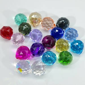 May include: A collection of faceted crystal balls in various colors, including red, pink, purple, blue, green, yellow, and clear. The spheres have a cut-glass appearance, reflecting light and creating a sparkling effect. The arrangement is scattered on a white surface.