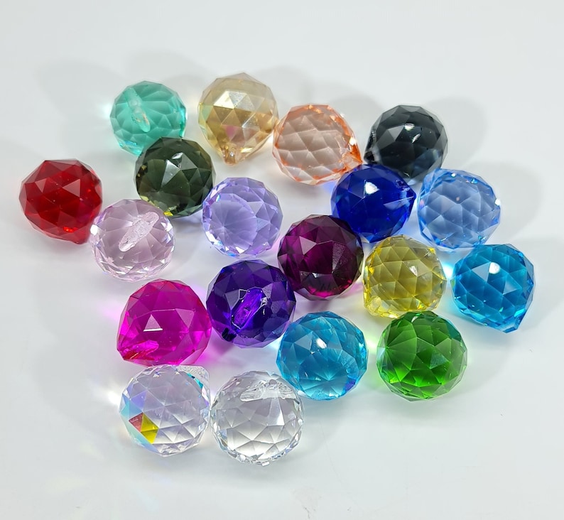 May include: A collection of faceted crystal balls in various colors, including red, pink, purple, blue, green, yellow, and clear. The spheres have a cut-glass appearance, reflecting light and creating a sparkling effect. The arrangement is scattered on a white surface.