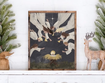 May include: A framed painting depicting a nativity scene with angels. The artwork features a dark background with white-robed angels surrounding a baby in a manger. The frame is made of dark wood. The painting is displayed on a white mantel.
