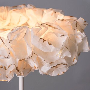Puede incluir: Una pantalla de lámpara de papel blanco con detalles dorados, que se asemeja a una flor, está unida a un soporte de metal blanco.