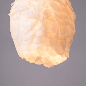 May include: A white paper lampshade with a unique, textured design that resembles overlapping scales. The lampshade is illuminated from within, casting a warm glow.