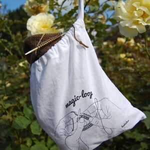 May include: White cotton drawstring bag with the text "magic-loop" and a black and white illustration of a hand knitting with a circular needle. The bag is hanging from a rose bush.