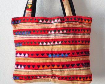 May include: A red, beige and blue tote bag with a geometric pattern. The bag has black straps and is hanging on a hook.