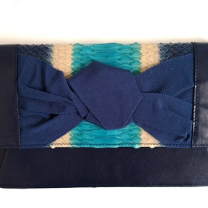Puede incluir: Clutch azul marino con detalle de lazo. El clutch presenta un estampado de piel de serpiente en tonos azules, turquesas y beige. El lazo es de tela azul marino a juego. Forma rectangular.