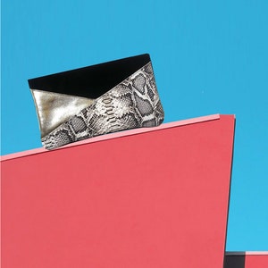Può includere: Una elegante pochette con motivo a pelle di serpente, pannelli metallici dorati e in camoscio nero. La borsa è presentata su uno sfondo rosso e blu vivace, che ne evidenzia il design e la consistenza.