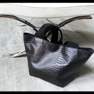 Peut inclure: Un sac fourre-tout noir avec un motif peau de serpent. Le sac a deux poignées noires et un dessus en daim. Le sac est posé sur une surface grise. Le sac est près d'une chaise blanche avec des pieds chromés.