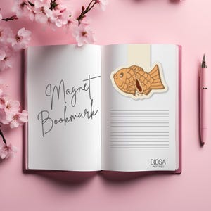 May include: A brown and tan fish-shaped magnet bookmark with a brown filling. The bookmark is on a white page with the text "Magnet Bookmark" written in black. The page is open in a notebook with a pink background.