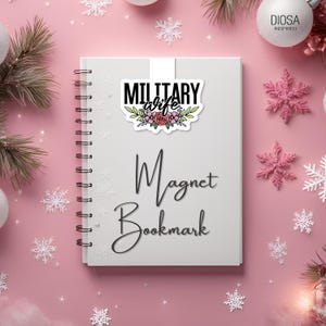 Op de afbeelding: Een witte magneet bladwijzer met de woorden "MILITARY wife" in het zwart, met bloemaccenten. De woorden "Magnet Bookmark" staan eronder in cursief. De achtergrond is roze met sneeuwvlok- en kerstversieringen.