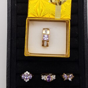 Puede incluir: Una caja de anillo de terciopelo negro con un anillo de oro con una piedra cuadrada púrpura y piedras más pequeñas transparentes. La caja está abierta y el anillo está dentro. Hay tres anillos más con piedras púrpuras en el fondo de la caja.