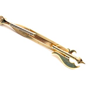 May include: A gold-toned tie clip with a decorative design. The clip features a central silver-colored, twisted bar and a stylized axe-like end with green accents. The clip is set against a white background.