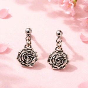 May include: A pair of silver-toned drop earrings. Each earring features a small ball stud connected to a chain, which leads to a detailed rose pendant. The background is a soft pink with flower petals.