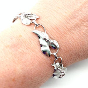 May include: A silver-toned bracelet featuring a leaf design. The bracelet has multiple leaf-shaped links connected by circular rings. Each leaf has a textured surface and small, round accents. The bracelet is worn on a wrist.