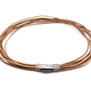 Op de afbeelding: Een meerstrengs ketting bestaande uit verschillende dunne, lichtbruine leren koorden in een cirkelvorm. De koorden worden bij elkaar gehouden door een rechthoekige, geborstelde zilverkleurige metalen sluiting met een zichtbaar scharnier en pin.