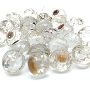 May include: A pile of clear quartz beads, each with multiple facets. The beads have internal inclusions of brown, red, and black. The beads are scattered on a white surface, creating a sparkling effect. Measurements are in centimetres.