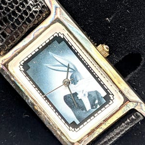 Pu&ograve; includere: Orologio da polso rettangolare con cassa color argento e cinturino in pelle nera. Il quadrante dell'orologio presenta un'immagine in bianco e nero di Bugs Bunny. L'orologio ha una corona dorata.