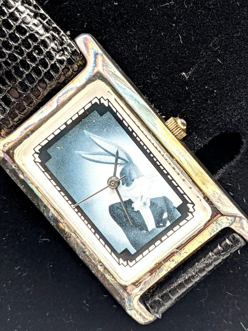 Pu&ograve; includere: Orologio da polso rettangolare con cassa color argento e cinturino in pelle nera. Il quadrante dell'orologio presenta un'immagine in bianco e nero di Bugs Bunny. L'orologio ha una corona dorata.