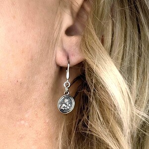 May include: A silver dangle earring with a circular charm featuring a detailed relief design. The earring is attached to a lever-back closure. The charm has a dark background with a detailed relief design.